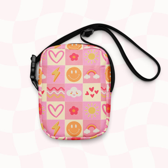 The Happy Smiles Sunny Days Mini Crossbody Bag by My Favourite Colour is Rainbow is a sturdy canvas bag decorated with bright icons like smiley faces, hearts, and more atop a pink checkered background.  It has internal and external pockets and a black strap with a clip on one side that can be extended to crossbody length.