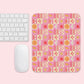 A computer keyboard and mouse on a pink surface. The mouse rests on the Happy Smiles Sunny Days Mouse Pad by My Favourite Colour is Rainbow, showcasing hearts, clouds, flowers, smiley faces, sun symbols, and lightning bolts in pink, yellow, orange, and red.