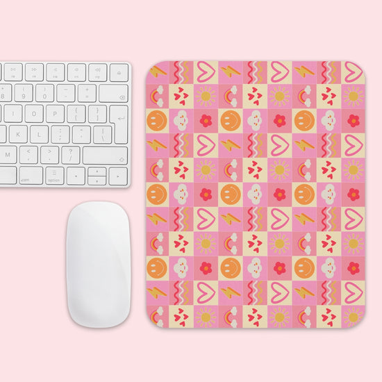 A computer keyboard and mouse on a pink surface. The mouse rests on the Happy Smiles Sunny Days Mouse Pad by My Favourite Colour is Rainbow, showcasing hearts, clouds, flowers, smiley faces, sun symbols, and lightning bolts in pink, yellow, orange, and red.