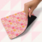 A hand lifts the corner of the Happy Smiles Sunny Days Mouse Pad by My Favourite Colour is Rainbow, showcasing a grid of cheerful icons like hearts, smiley faces, flowers, suns, and lightning bolts over a geometric pink and white patterned background.