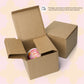 An open cardboard box displays the Happy Smiles Sunny Days Mug by My Favourite Colour is Rainbow, featuring a pink and orange smiley face design, beside a closed box. A note above ensures protection despite delivery variations. The background has a soft pink and cream pattern, enhancing the joyful vibe.