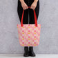 A person carries the stylish Happy Smiles Sunny Days Tote Bag by My Favourite Colour is Rainbow, featuring a red handle and a pattern of pink, yellow, and orange smiley faces, hearts, and abstract shapes. It complements their black pants and boots against a grey background.