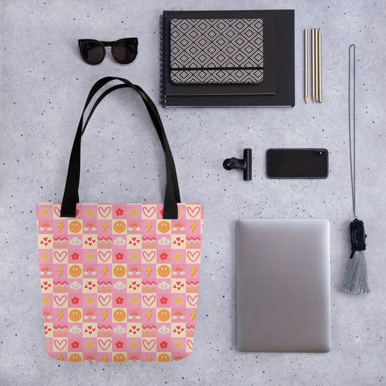 Flat lay featuring the Happy Smiles Sunny Days Tote Bag by My Favourite Colour is Rainbow, adorned with colorful emojis, alongside a laptop, smartphone, sunglasses, notebook, pen, and a tassel on a textured surface. 