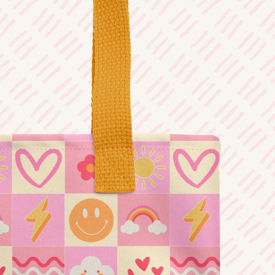 A close-up of the Happy Smiles Sunny Days Tote Bag by My Favourite Colour is Rainbow, showing the yellow handle.  The bag features a pink and cream checkerboard design with pink, red and yellow hearts, flowers, suns, lightning bolts, smiley faces, rainbows, clouds.