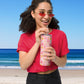 A person in a red shirt and sunglasses drinks from the My Favourite Colour is Rainbows Happy Smiles Sunny Days Stainless Steel Tumbler with a metal straw at the beach, framed by the ocean and clear blue sky on a sunny day.