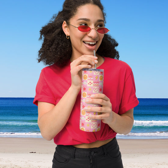 A person in a red shirt and sunglasses drinks from the My Favourite Colour is Rainbows Happy Smiles Sunny Days Stainless Steel Tumbler with a metal straw at the beach, framed by the ocean and clear blue sky on a sunny day.