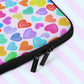 A close up of teh Heart Attack Laptop Sleeve by My Favourite Colour is Rainbow, featuring a print of colourful hearts. It has two black zippers. The print is on only one side of the laptop sleeve, the other side is black.  It rests on a pastel pink and blue striped background.