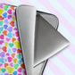 A My Favourite Colour is Rainbow Heart Attack Laptop Sleeve with a silver laptop partially pulled out reveals its vibrant heart-patterned design against a grey faux-fur interior. Shown on a soft pink and blue striped background.