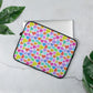 The 13" Heart Attack Laptop Sleeve by My Favourite Colour is Rainbow features a vibrant heart pattern in pink, blue, green, yellow, and orange. It sits on a light grey surface surrounded by green leaves and partially reveals a laptop inside.  The print is on only one side of the laptop sleeve, the other side is black.