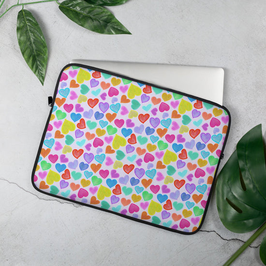 The 15" Heart Attack Laptop Sleeve by My Favourite Colour is Rainbow features a vibrant heart pattern in pink, blue, green, yellow, and orange. It sits on a light grey surface surrounded by green leaves and partially reveals a laptop inside.  The print is on only one side of the laptop sleeve, the other side is black.