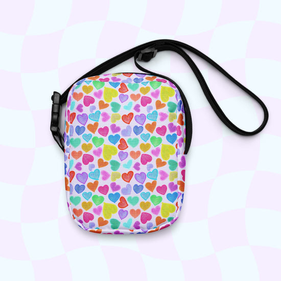 The Heart Attack Mini Crossbody Bag by My Favourite Colour is Rainbow has adjustable straps and a vibrant Heart Attack print with assorted bright hearts on a white background, set against a light checkered backdrop.