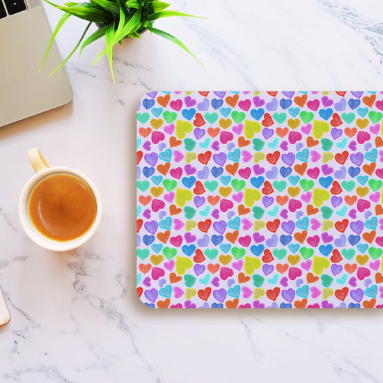 A vibrant Heart Attack Mouse Pad by My Favourite Colour is Rainbow features multicolored hearts on a marble design. Near it are a laptop, coffee cup, and green plant, creating a bright, cheerful workspace.