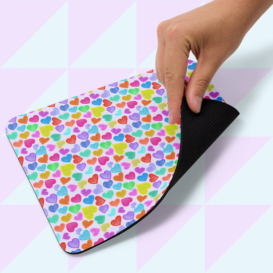 A hand lifts the corner of the Heart Attack Mouse Pad by My Favourite Colour is Rainbow, featuring multicolored hearts on a light background. The pads underside is black, and it rests on a surface with a pastel geometric design.