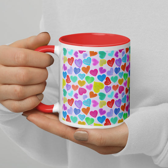 A person in a white shirt holds the Heart Attack Mug by My Favourite Colour is Rainbow, featuring a red interior and handle with a vibrant, multicoloured heart pattern.