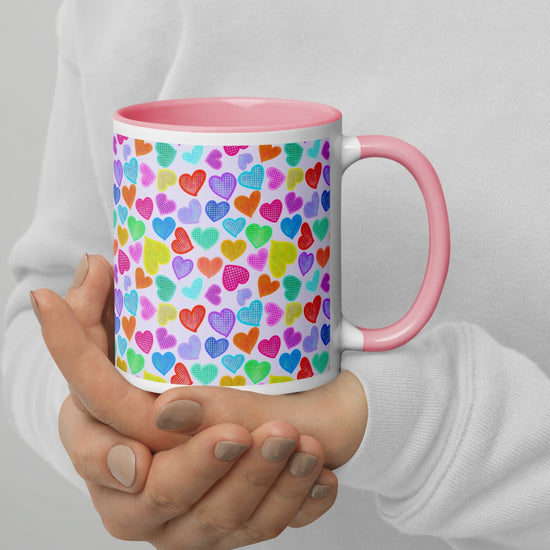 A person wearing a white sweater holds the Heart Attack Mug from My Favourite Colour is Rainbow, featuring a pink interior and handle, with a heart pattern in shades of blue, red, green, yellow, and purple.