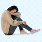 A person sits on the ground in a comfy fit, wearing a beige sweater, dark pants, and colourful Heart Attack Canvas Sneakers by My Favourite Colour is Rainbow. They rest their head on their knee, looking relaxed.