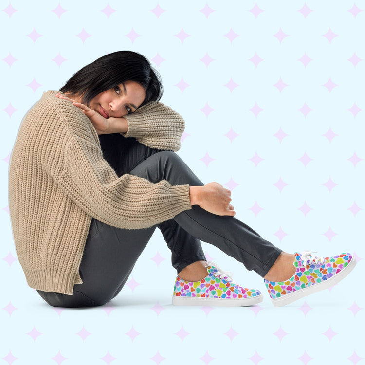 A person sits on the ground in a comfy fit, wearing a beige sweater, dark pants, and colourful Heart Attack Canvas Sneakers by My Favourite Colour is Rainbow. They rest their head on their knee, looking relaxed.