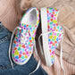 The Heart Attack Canvas Sneakers by My Favourite Colour is Rainbow feature a vibrant heart pattern in blue, purple, pink, orange, and green on a light background. With white laces and soles for comfort, they're styled on light pink jumper next to folded denim jeans.