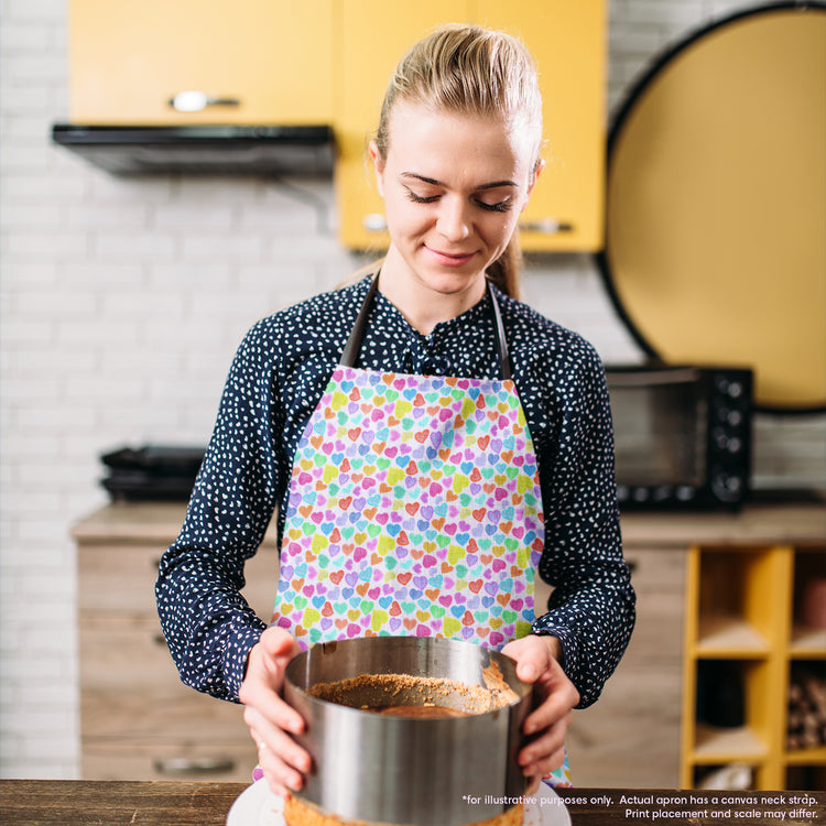 In a modern kitchen with yellow cabinets, a woman wearing a vibrant Heart Attack Apron by My Favourite Colour is Rainbow holds a round cake pan. Focused on her culinary task, she works diligently with various kitchen appliances visible in the background.  The apron features a colourful heart pattern on a light background.