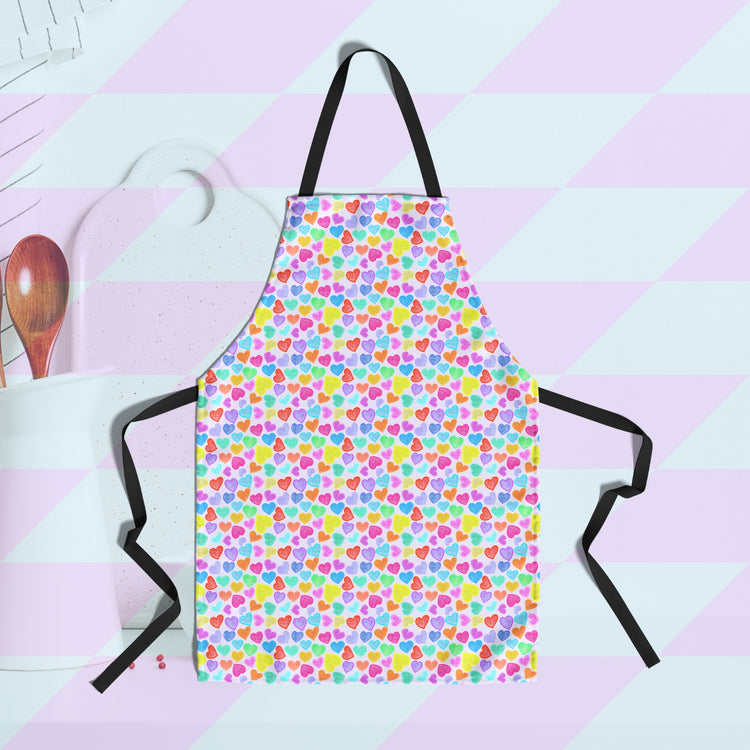 Displayed is the Heart Attack Apron by My Favourite Colour is Rainbow, featuring a colourful heart pattern on a light background with black neck and waist ties. In the background, Kitchen utensils are partially visible to the left in the background.