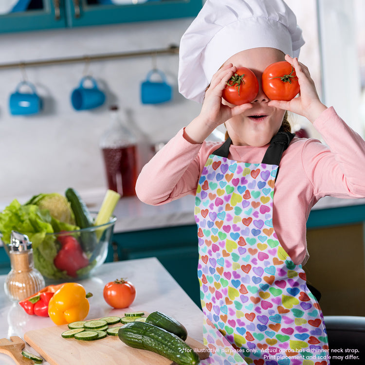 A child in a chefs hat and a Heart Attack Apron by My Favourite Colour is Rainbow playfully holds two tomatoes over their eyes. The kitchen counter, ready for an exciting cooking adventure, showcases colourful vegetables like cucumbers, tomatoes, and leafy greens.  The apron features a colourful heart pattern on a light background.