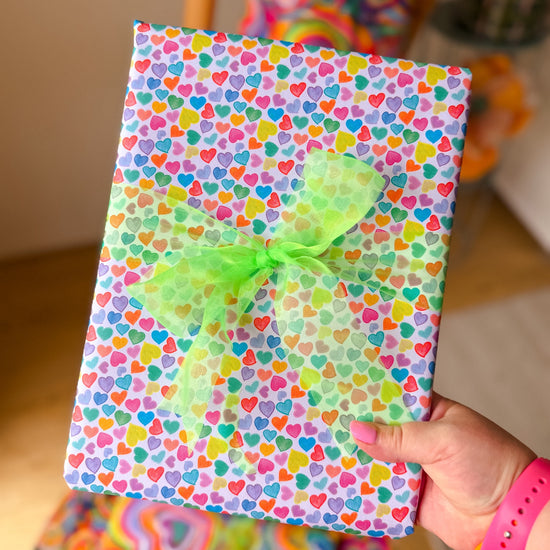 A hand holds a gift wrapped in Heart Attack Wrapping Paper Sheets by My Favourite Colour is Rainbow, with a vibrant heart design in blue, pink, yellow, and green. The eco-friendly gift is topped with a light green translucent bow and features a hint of a rainbow background for added charm.