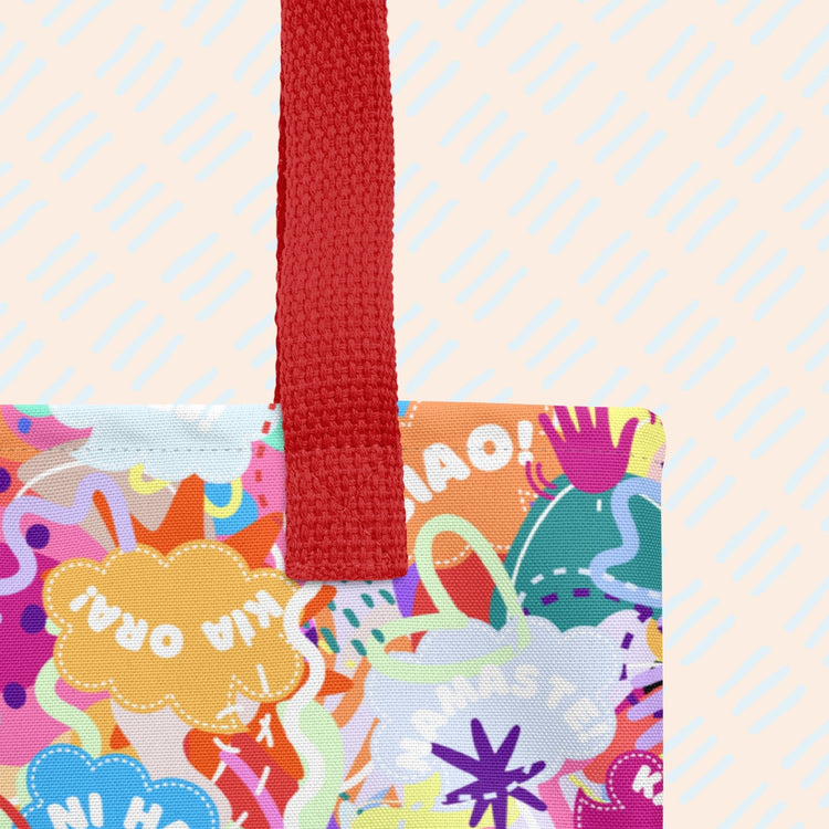 A close-up of The Hello! Wominjeka! Tote Bag by My Favourite Colour is Rainbow showing the sturdy red strap and bold abstract designs with "Hello" in various languages like hola and bonjour. 