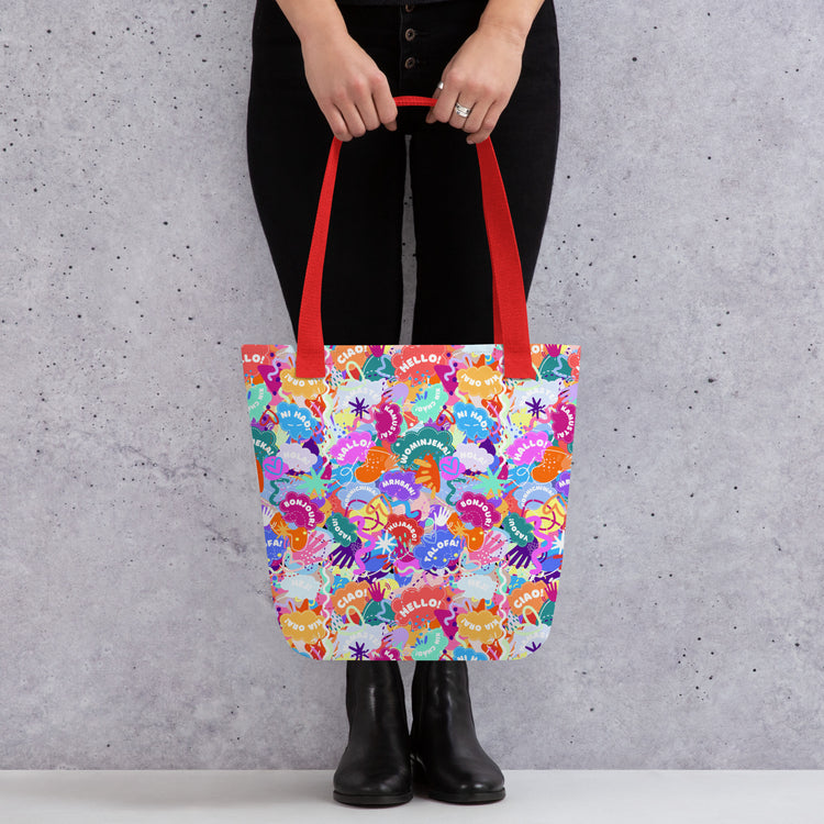 Someone is holding the Hello! Wominjeka! Tote Bag by My Favourite Colour is Rainbow, featuring a bold abstract design with "Hello" in various languages.  The bag has red handles, contrasting with their black pants and boots.