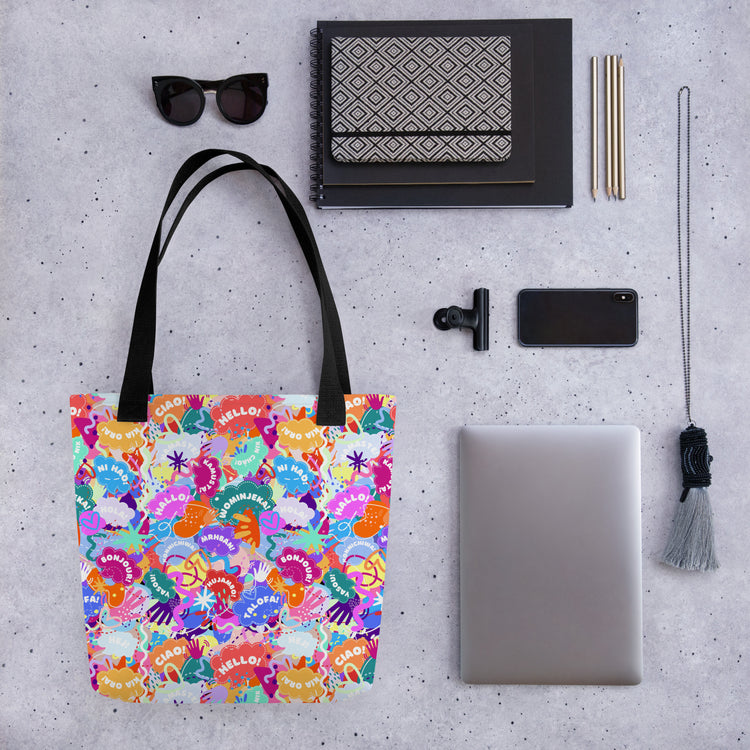 A flat lay of the stylish Hello! Wominjeka! Tote Bag from My Favourite Colour is Rainbow, featuring a bold abstract design with "Hello" in various languages.. Nearby are sunglasses, notebooks, pens, a smartphone, clip, laptop, and tassel keychain.