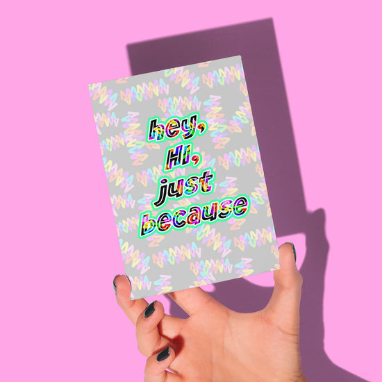 A hand with dark nail polish holds My Stores eco-friendly Hey, Hi, Just Because Greeting Card against a pink background. Featuring a colorful, wavy pattern with bold multicolored text, its shadow is visible on the background.