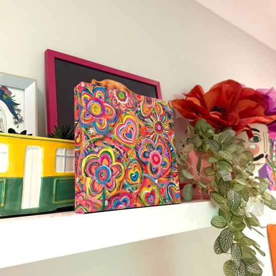 The Love in Neon canvas artwork by My Favourite Colour is Rainbow, featuring colorful hearts and swirls, graces the shelf amidst vibrant décor: a ceramic yellow bus, a plant, a large red flower, and framed items against a light-colored wall.