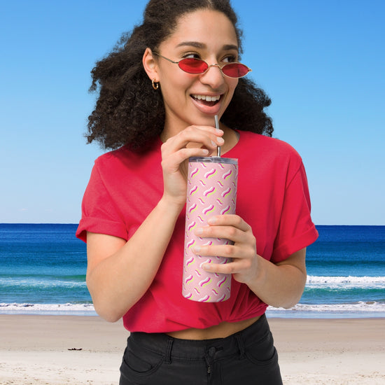 A woman in a red shirt and small red sunglasses smiles, sipping from the Iced Vovo Stainless Steel Tumbler by My Favourite Colour is Rainbow, featuring a pink pattern and metal straw. She stands in front of a beach with blue sky and ocean waves in the background.