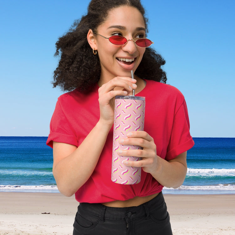 A woman in a red shirt and small red sunglasses smiles, sipping from the Iced Vovo Stainless Steel Tumbler by My Favourite Colour is Rainbow, featuring a pink pattern and metal straw. She stands in front of a beach with blue sky and ocean waves in the background.