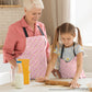 A woman and a young girl are baking together in matching Iced Vovo Rainbow Aprons from My Favourite Colour is Rainbow. The girl skillfully rolls dough, surrounded by milk and flour containers. The Iced Vovo aprons feature an abstract pink, white and green design reminiscent of the famous biscuit.