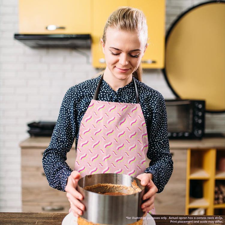 Wearing the Iced Vovo Rainbow Apron by My Favourite Colour is Rainbow, a woman lifts a round silver baking pan to reveal a cake. She concentrates on her task, surrounded by kitchen cabinets, an oven, and a microwave in the background.  The Iced Vovo apron features an abstract pink, white and green design reminiscent of the famous biscuit.