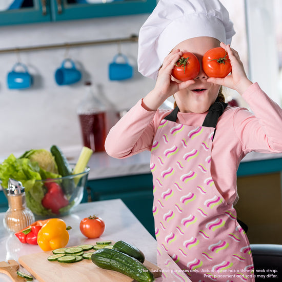 A child in a chef hat and My Favourite Colour is Rainbows Iced Vovo Rainbow Apron playfully holds tomatoes over their eyes in a kitchen, showcasing their culinary creativity. Fresh vegetables like cucumbers, bell peppers, and lettuce decorate the countertop before them.  The Iced Vovo apron features an abstract pink, white and green design reminiscent of the famous biscuit.