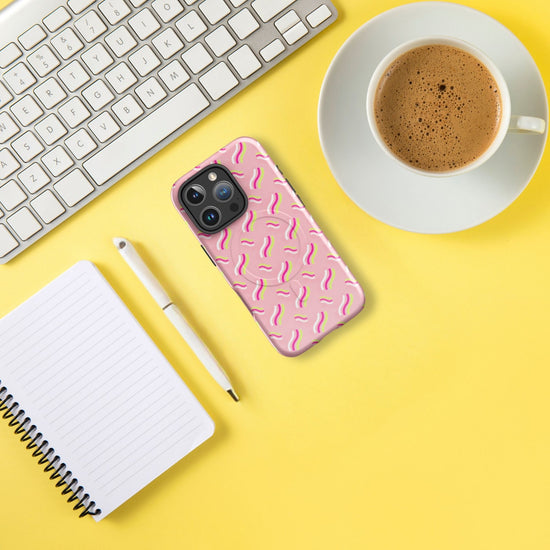 An iPhone with an Iced Vovo MagSafe case by My Favourite Colour is Rainbow, a coffee cup on a saucer, a white pen, a spiral notebook with lined pages, and a wireless keyboard are displayed on a bright yellow surface.