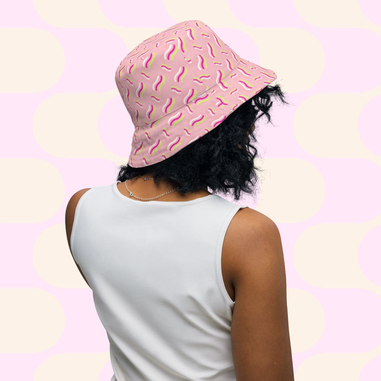 Sporting curly hair, a person wears the My Favourite Colour is Rainbows In the Midnight Garden/Iced Vovo pink reversible bucket hat with a squiggle pattern and a white sleeveless top against a pink and cream geometric backdrop—ideal for anyone in search of their Style BFF.