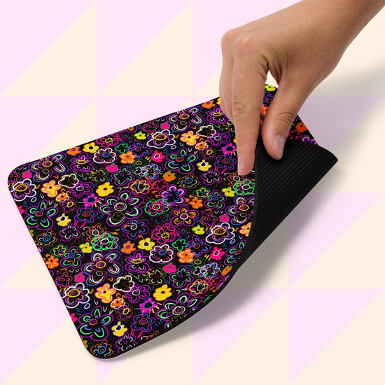 A hand lifts a corner of the In the Midnight Garden Mouse Pad from My Favourite Colour is Rainbow, featuring a vibrant floral pattern on a black background with a solid black underside. The background displays light pink and cream triangular shapes.