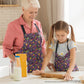 A woman and a young girl enjoy cooking together wearing matching In the Midnight Garden aprons by My Favourite Colour is Rainbow. The girl skillfully rolls dough while milk and a flour box are on the table.  The aprons feature a striking design of multicoloured flowers on a black background.  