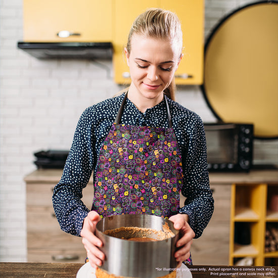 Someone wearing an In the Midnight Garden apron by My Favourite Colour is Rainbow holds a baking pan with a crust inside, standing in a kitchen featuring yellow cabinets, a microwave, and a stand mixer.  The apron features a striking design of multicoloured flowers on a black background.  