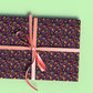 A gift wrapped in In the Midnight Garden Wrapping Paper by My Favourite Colour is Rainbow, features a floral and neon flower pattern on a dark background.  The gift is tied with a pink ribbon.