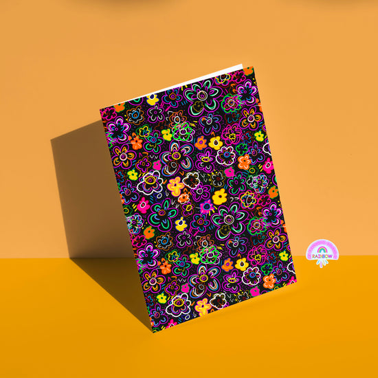 The In the Midnight Garden Greeting Card by My Store features neon floral patterns on a black background akin to a midnight garden, casting a shadow on a tan surface with a rainbow logo in the bottom right.