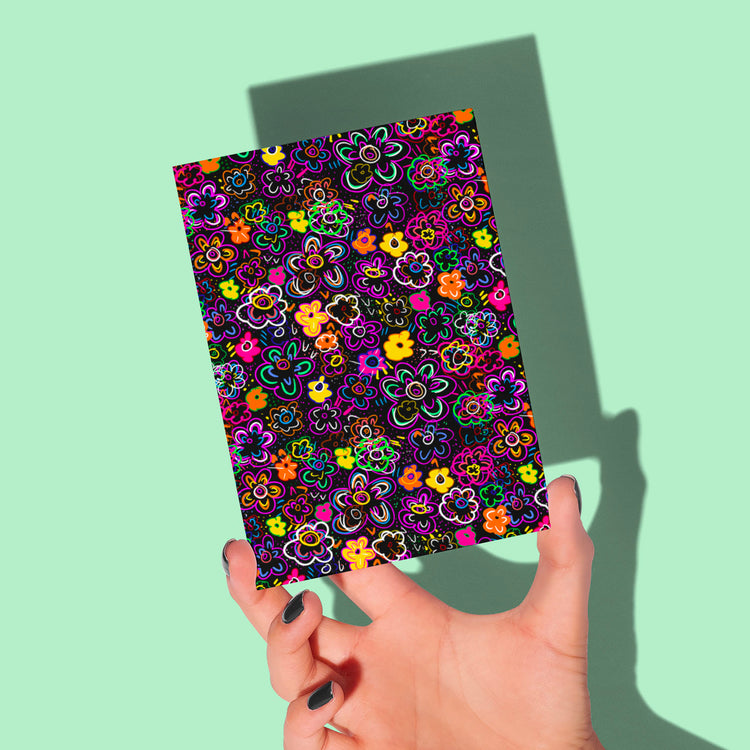 A hand with dark nail polish holds the In the Midnight Garden Greeting Card from My Store, showcasing neon blooms on a detailed floral black background, casting a shadow on a mint green surface.