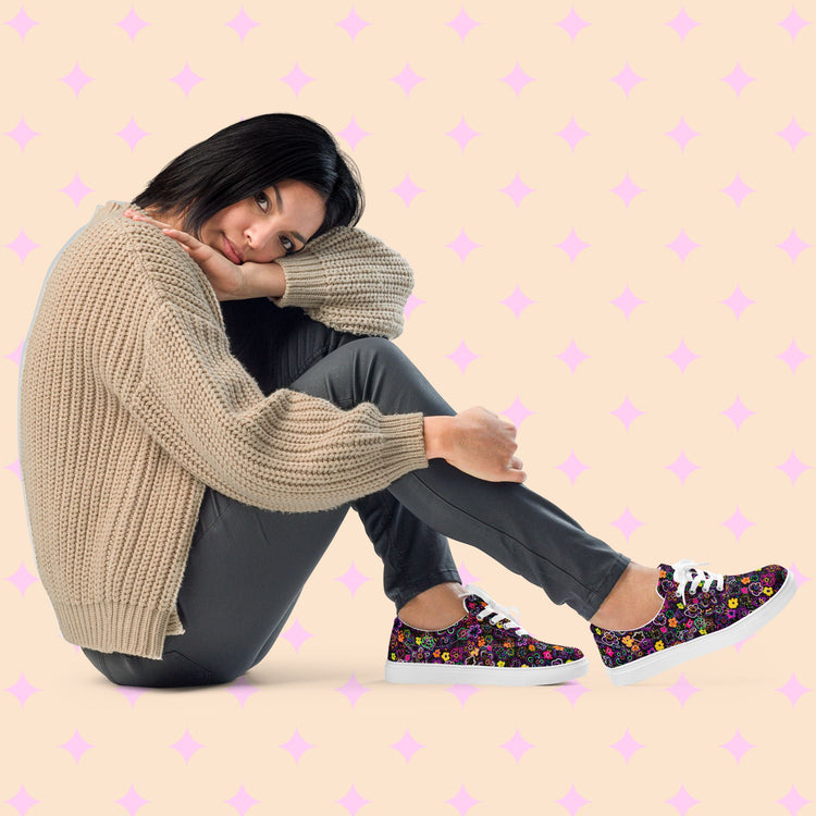 A person with shoulder-length hair sits on the floor, leaning their head on their knee. They're dressed in a beige knitted sweater, grey pants, and My Favourite Colour is Rainbow's In the Midnight Garden canvas sneakers. The background features a pink diamond pattern on light peach.