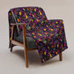 A wooden armchair with a gray cushion displays the In the Midnight Garden Throw Blanket by My Favourite Colour is Rainbow, featuring vibrant skull and flower patterns on a dark background. The chair sits on a neutral-toned floor.