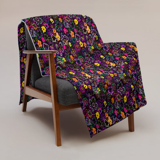 A wooden armchair with a gray cushion displays the In the Midnight Garden Throw Blanket by My Favourite Colour is Rainbow, featuring vibrant skull and flower patterns on a dark background. The chair sits on a neutral-toned floor.