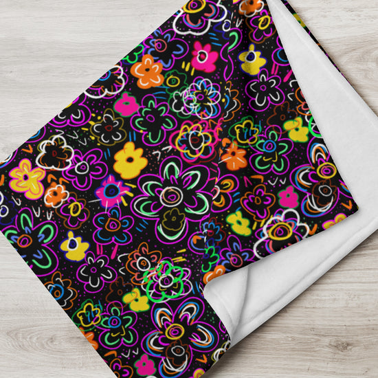 The In the Midnight Garden Throw Blanket by My Favourite Colour is Rainbow features abstract neon pink, yellow, orange, and green floral patterns on a black background. Crafted from cozy fabric, it looks perfect on a light wooden surface.