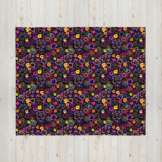 The In the Midnight Garden Throw Blanket by My Favourite Colour is Rainbow showcases a vibrant, cozy fabric with multicolored flowers in shades of purple, orange, yellow, red, and blue on a dark background—ideal for styling on a light wood surface.