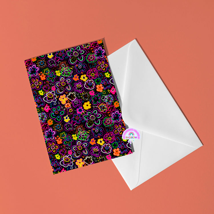 The In the Midnight Garden Greeting Card by My Store features a neon floral pattern partially tucked in a white envelope against a coral background, enhanced with a small rainbow-themed sticker.